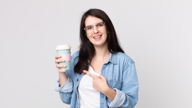 Jolie femme souriante joyeusement, se sentant heureuse et pointant sur le côté et tenant un café à emporter