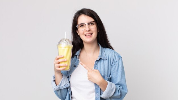 Jolie femme souriante joyeusement, se sentant heureuse et pointant sur le côté et tenant un milk-shake à la vanille