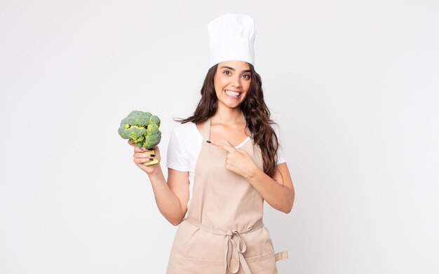 Jolie femme souriante joyeusement, se sentant heureuse et pointant vers le côté portant un tablier et tenant un brocoli