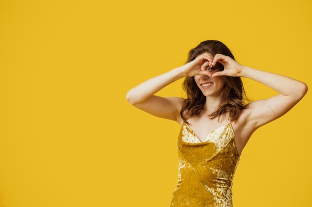 Jolie femme souriante montrant le signe du coeur avec les mains près des yeux posant en robe sur l'espace libre de fond jaune