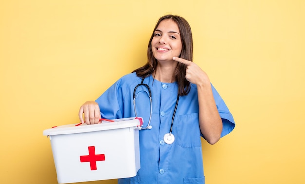 Jolie femme souriante pointant avec confiance vers son large sourire. infirmière avec une trousse de premiers soins infirmière