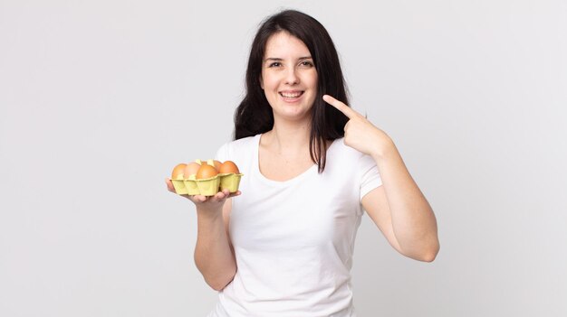 Jolie femme souriante pointant avec confiance vers son large sourire et tenant une boîte à œufs