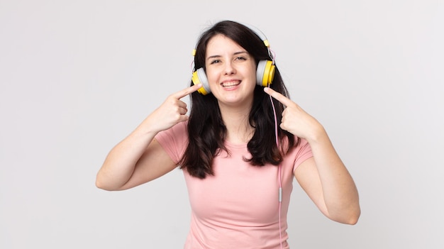 Jolie femme souriante pointant du doigt son propre large sourire en écoutant de la musique avec des écouteurs