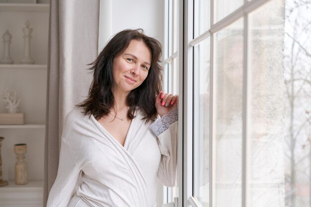 Jolie femme souriante rêvassant debout à la fenêtre en peignoir Parmi