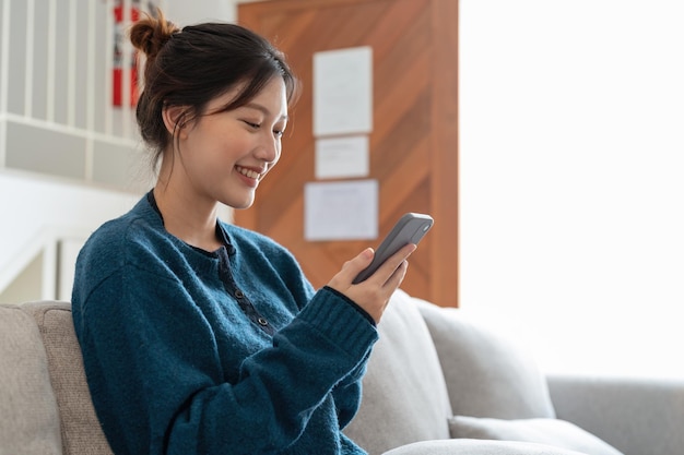 Jolie femme souriante utilisant un téléphone intelligent assis sur le canapé à la maison Concept de communication et de confort