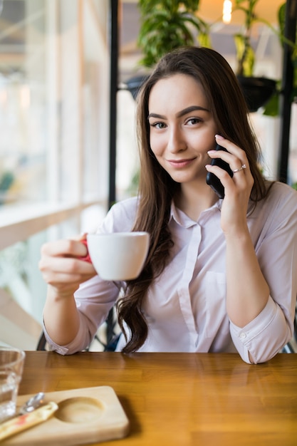 Jolie femme avec un sourire mignon ayant parler de conversation avec un téléphone mobile tout en restant au café