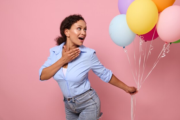 Jolie femme surprise joyeuse d'ethnie métisse regarde des ballons à air lumineux multicolores dans ses mains, isolées sur fond rose avec espace de copie pour le texte et la publicité