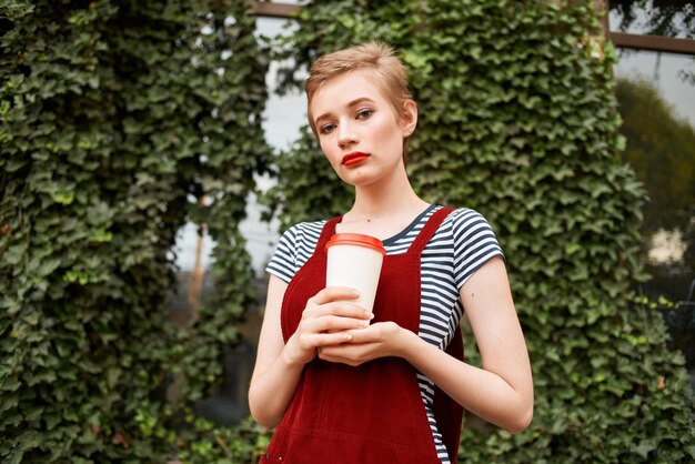 Jolie femme avec une tasse de boisson dans la rue près des buissons