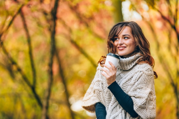 Jolie femme avec une tasse de café sourit et regarde ailleurs dans le parc à l'automne.