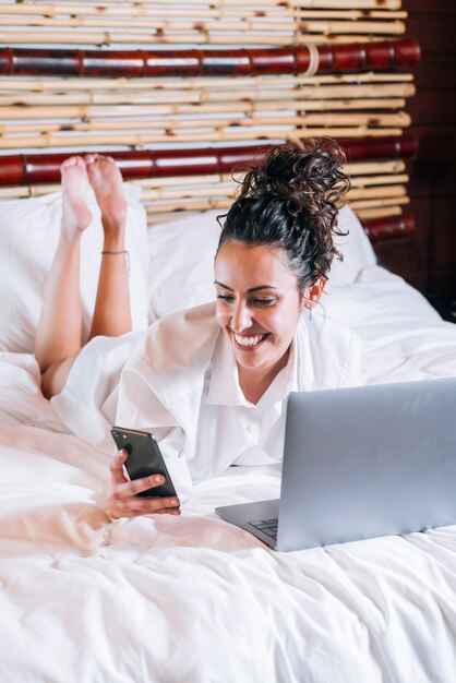 Jolie femme avec téléphone et ordinateur portable au lit