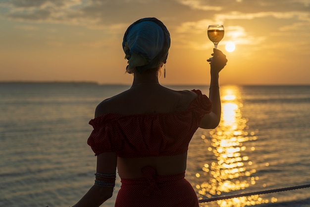 Jolie femme tenant un verre de vin blanc contre un beau coucher de soleil près des vagues de la mer sur la plage tropicale, gros plan. La fille profite du coucher de soleil avec un verre de vin. Concept de loisirs et de voyage