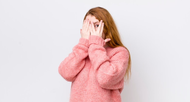 Jolie femme à tête rouge couvrant le visage avec les mains furtivement entre les doigts avec une expression surprise et regardant sur le côté