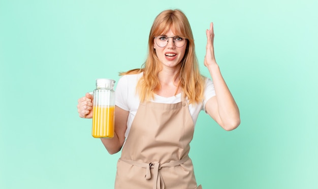 Photo jolie femme à tête rouge criant avec les mains en l'air avec un tablier préparant un jus d'orange