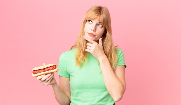 Jolie femme à tête rouge pensant, se sentant dubitative et confuse et tenant un hot-dog