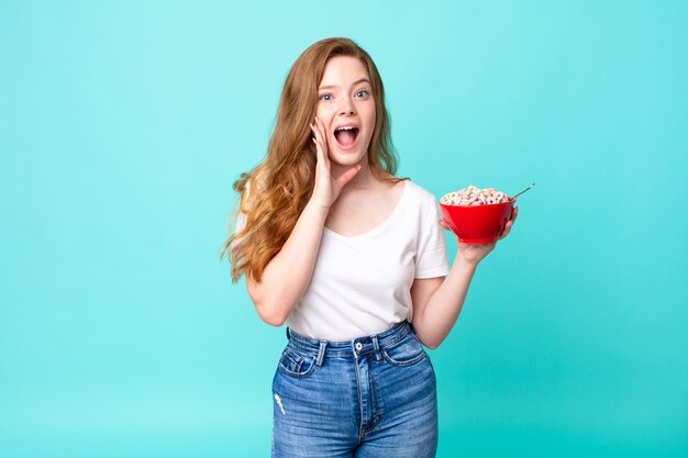 Jolie femme à tête rouge se sentant choquée et effrayée et tenant un bol de petit-déjeuner