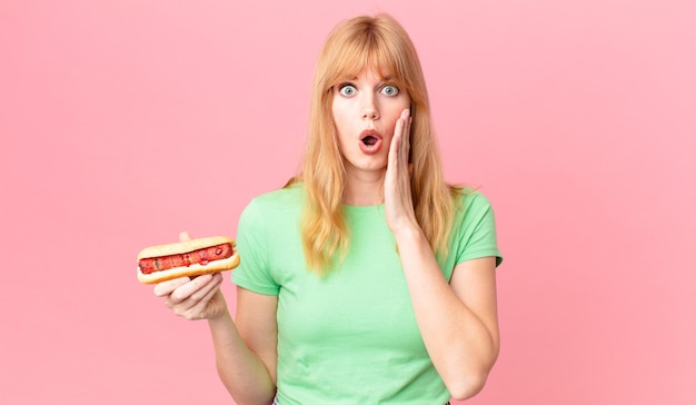 Jolie femme à tête rouge se sentant choquée et effrayée et tenant un hot-dog