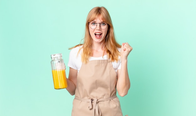 Jolie femme à tête rouge se sentant choquée, riant et célébrant le succès avec un tablier préparant un jus d'orange