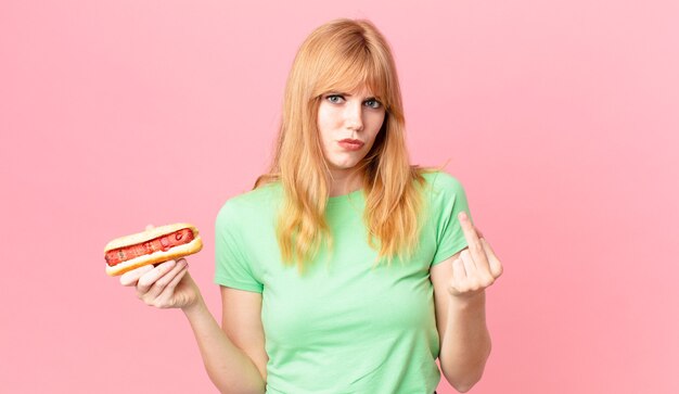 Photo jolie femme à tête rouge se sentant en colère, agacée, rebelle et agressive et tenant un hot-dog