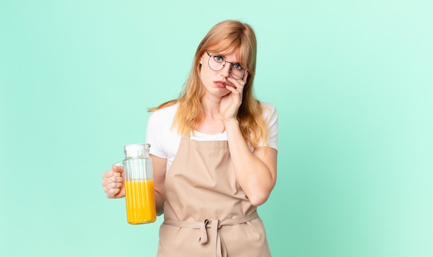 Jolie femme à tête rouge se sentant ennuyée, frustrée et somnolente après un fastidieux avec un tablier préparant un jus d'orange