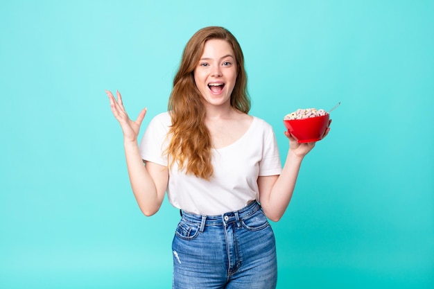 Jolie femme à tête rouge se sentant heureuse et étonnée de quelque chose d'incroyable et tenant un bol de petit-déjeuner