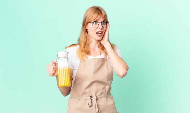 Jolie femme à tête rouge se sentant heureuse, excitée et surprise avec un tablier préparant un jus d'orange