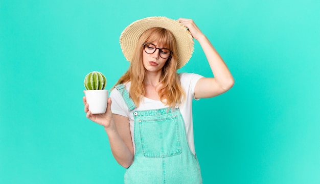 Jolie femme à tête rouge se sentant perplexe et confuse, se grattant la tête et tenant un cactus en pot. concept d'agriculteur