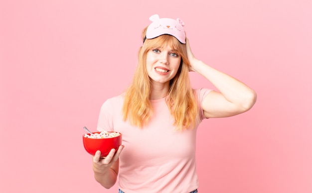 Jolie femme à tête rouge se sentant stressée, anxieuse ou effrayée, les mains sur la tête en pyjama et tenant un bol de flocons