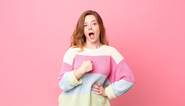 Jolie femme à tête rouge semblant choquée et surprise avec la bouche grande ouverte, pointant vers soi