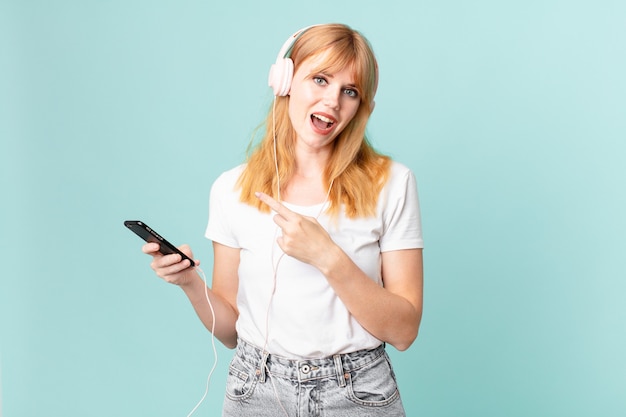 Jolie femme à tête rouge semblant excitée et surprise en pointant sur le côté et en écoutant de la musique avec des écouteurs