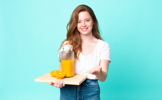 Jolie femme à tête rouge souriant joyeusement avec amicale et offrant et montrant un concept et tenant un jus d'orange