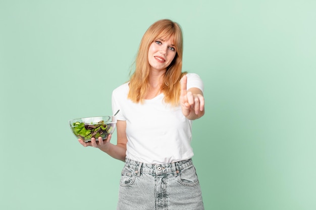 Jolie femme à tête rouge souriante fièrement et avec confiance faisant le numéro un et tenant une salade. concept de régime