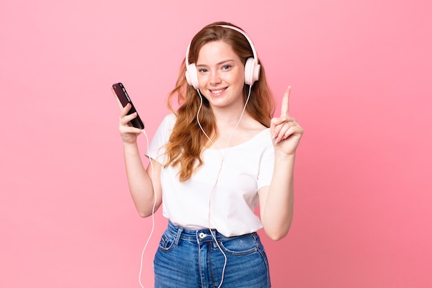 Jolie femme à tête rouge souriante et semblant amicale, montrant le numéro un avec un casque et un smartphone