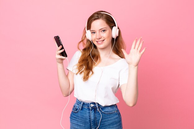 Jolie femme à tête rouge souriante et semblant amicale, montrant le numéro cinq avec un casque et un smartphone