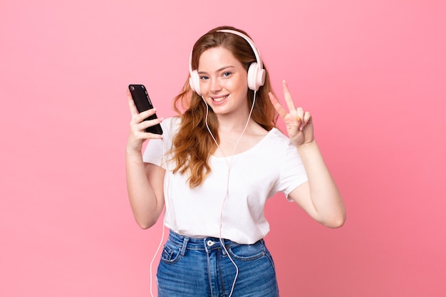 Jolie femme à tête rouge souriante et semblant heureuse, gesticulant la victoire ou la paix avec des écouteurs et un smartphone