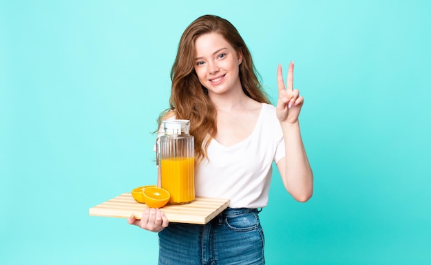 Jolie femme à tête rouge souriante et semblant heureuse, gesticulant la victoire ou la paix et tenant un jus d'orange