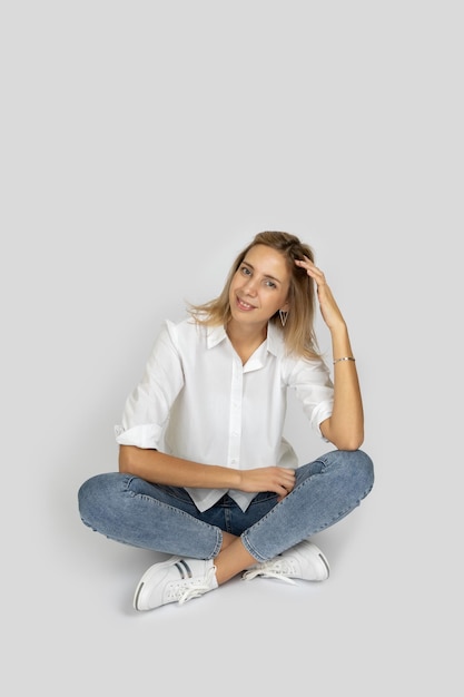 Jolie femme touchant les cheveux avec la main posant assis avec les jambes croisées sur fond de studio blanc regardant la caméra en chemisier blanc jeans et baskets montrant des émotions positives