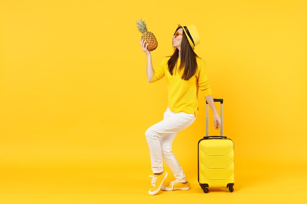 Jolie femme touristique voyageuse en vêtements d'été, chapeau tenant des fruits frais d'ananas mûrs isolés sur fond orange jaune. Passager voyageant à l'étranger en week-end. Concept de voyage en avion