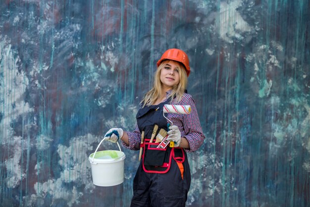 Jolie femme en uniforme et casque tenant de la peinture et un rouleau se préparant à rénover la maison. Ouvrier du batiment.