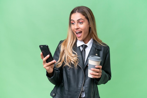 Jolie femme uruguayenne d'affaires portant une veste de motard sur fond isolé tenant du café à emporter et un mobile