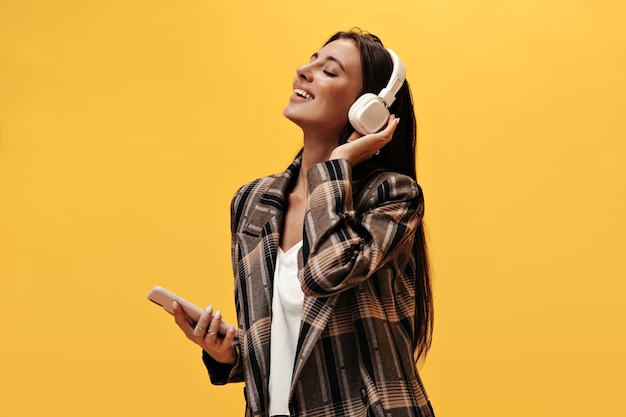 Jolie femme en veste marron élégante et tee blanc tient le téléphone et sourit Jolie fille écoutant de la musique dans les écouteurs sur fond jaune