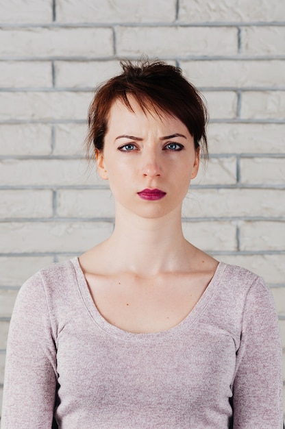 Une jolie femme avec un visage fronçant les sourcils, de grands yeux bleus, des lèvres roses pleines, un mur de briques blanches