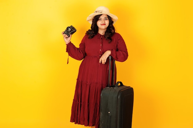 Jolie femme de voyage sud-asiatique en robe rouge foncé robe chapeau posé au studio sur fond jaune avec valise amd vieil appareil photo vintage