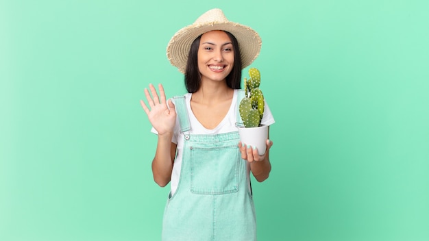 jolie fermière souriante joyeusement, agitant la main, vous accueillant et vous saluant et tenant un cactus