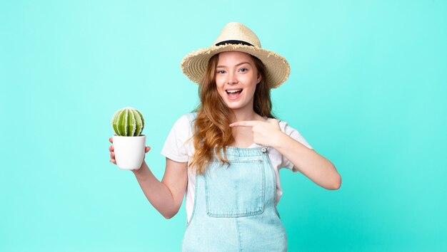 Jolie fermière à tête rouge à la recherche d'excitation et de surprise pointant sur le côté et tenant un cactus