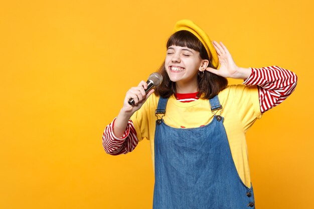 Jolie fille adolescente en béret français, robe d'été en denim gardant les yeux fermés chante une chanson dans un microphone isolé sur fond jaune en studio. Concept de mode de vie des émotions sincères des gens. Maquette de l'espace de copie.