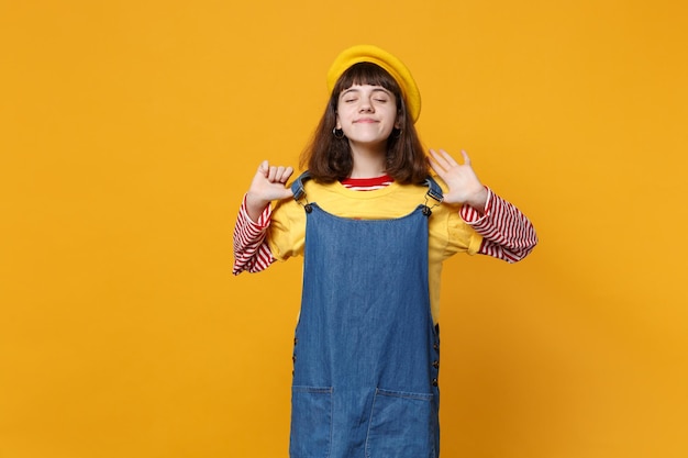 Jolie fille adolescente en béret français tenant la main sur la robe d'été en denim, gardant les yeux fermés isolés sur fond de mur jaune en studio. Émotions sincères des gens, concept de style de vie. Maquette de l'espace de copie.