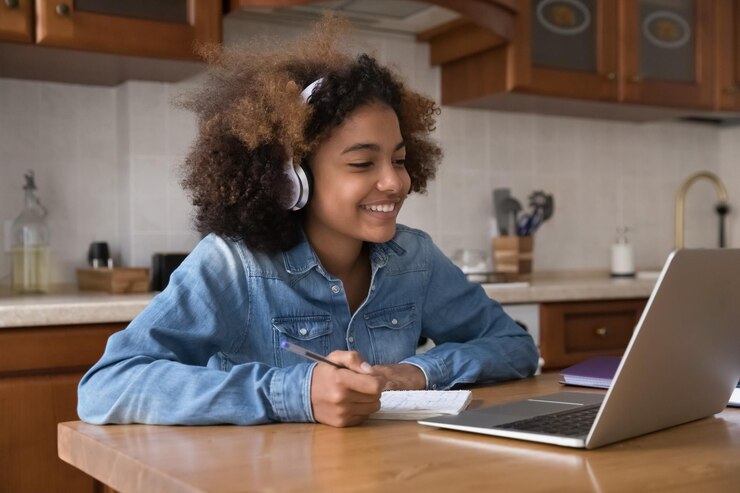 Photo une jolie fille africaine porte des écouteurs, étudie, s'assoit à la table dans la cuisine, utilise un ordinateur portable, s'engage dans une classe distante avec un tuteur en ligne, sourit, apprécie l'apprentissage, se sent motivée.