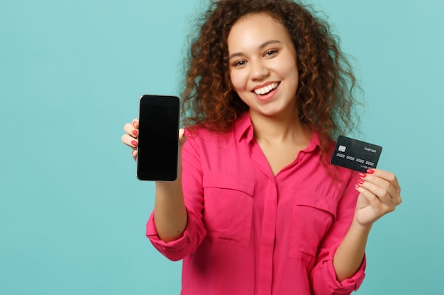 Une jolie fille africaine en vêtements décontractés tient un téléphone portable avec un écran vide vierge, une carte bancaire de crédit isolée sur fond bleu turquoise. Les gens émotions sincères, concept de style de vie. Maquette de l'espace de copie.