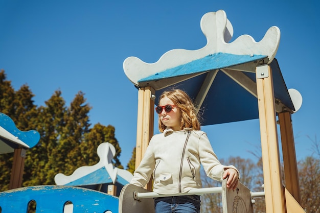 Photo jolie fille d'âge préscolaire grimpant sur un équipement de terrain de jeu sur fond de ciel bleu, une fille heureuse en veste beige et des lunettes de soleil joue sur une aire de jeux en plein air