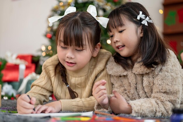 Une jolie fille asiatique aide sa sœur à fabriquer une carte de Noël en étant allongée sur le sol ensemble